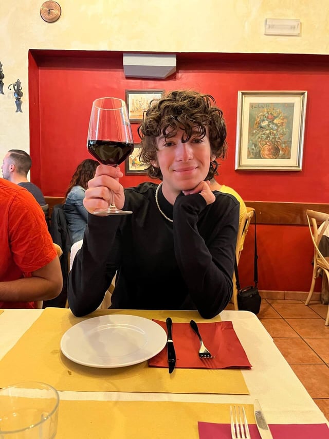 An individual wearing a black turtleneck holding up a glass of wine while seated in a restaurant