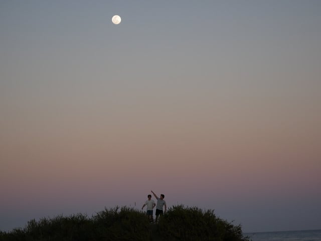 Two students atop a hill pointing to the Moon in the Dunes of Maspalomas