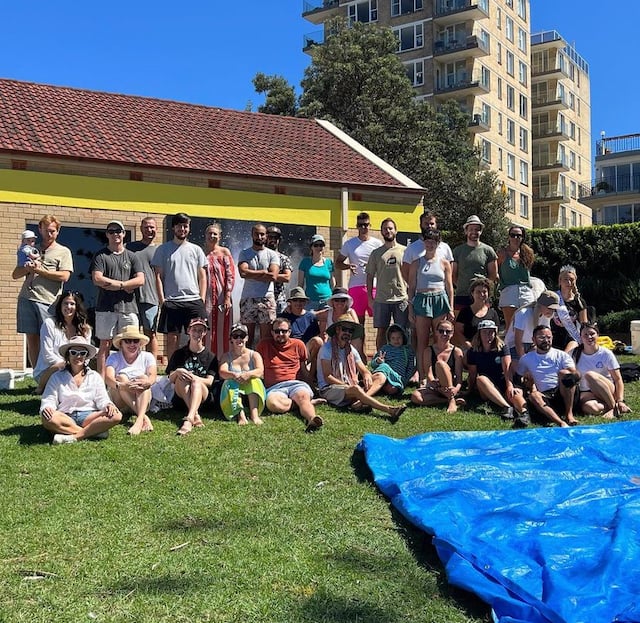 A group photo of people standing and sitting on the grass after a beach cleanup with a blue plastic sheet in front of them