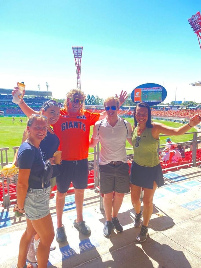 Five people pose happily at a GWS GIANTS game in Sydney with the field and stadium in the background